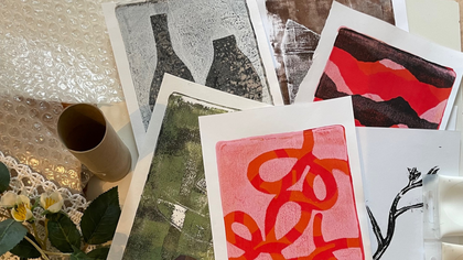 Flera abstrakta konsttryck i olika färger ligger utspridda på ett bord med blommor och bubbelplast.