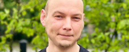 En man med rakat huvud och svart t-shirt med texten 'musik' står utomhus framför gröna buskar.