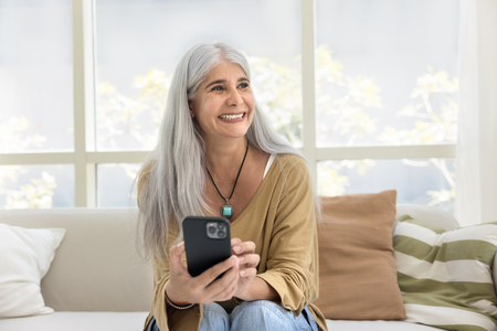 Kvinna med långt grått hår sitter i soffa och håller en mobiltelefon i vardagsrum med stora fönster.