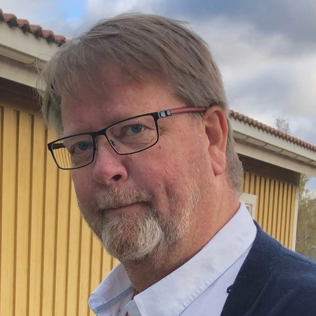 Man med glasögon och skägg står utomhus framför ett gult trähus under molnig himmel.