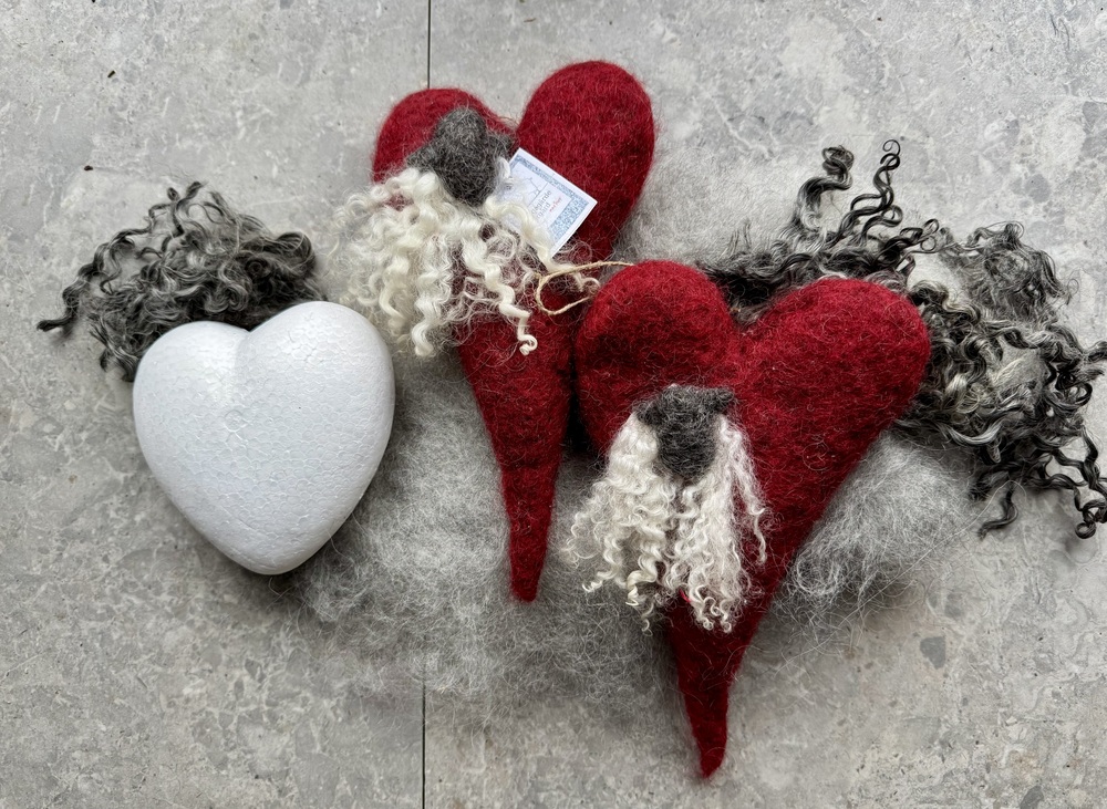 Två röda hjärtformade ullfigurer med skägg och en vit hjärtformad dekoration på grått underlag.