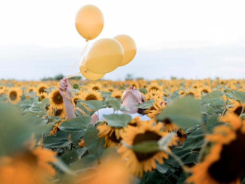 Person håller tre gula ballonger i ett fält av solrosor under en ljus himmel.