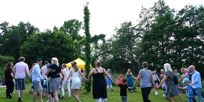 En grupp människor dansar i ring utomhus runt en midsommarstång på en gräsmatta.