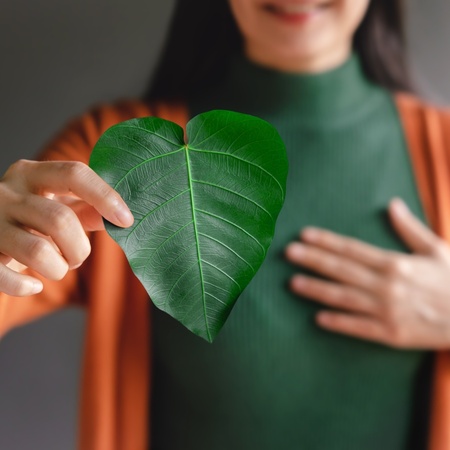 Kvinna håller ett grönt hjärtformat blad framför bröstet, klädd i grön tröja och orange kofta.