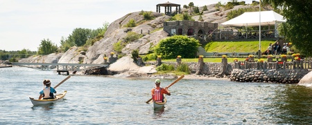 Två personer paddlar kanot på en sjö nära en klippig strand med grönska och byggnader.