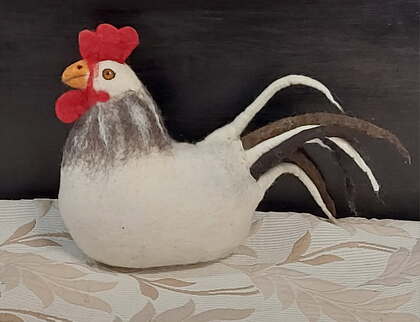 En filtad tupp i vitt, rött och brunt står på ett mönstrat tyg framför en mörk bakgrund.