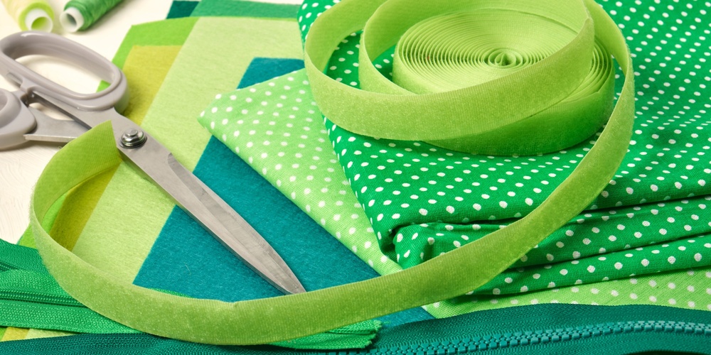 Sytillbehör i gröna nyanser, inklusive tyg, sax, dragkedja, tråd och band, ligger utspridda på ett bord.