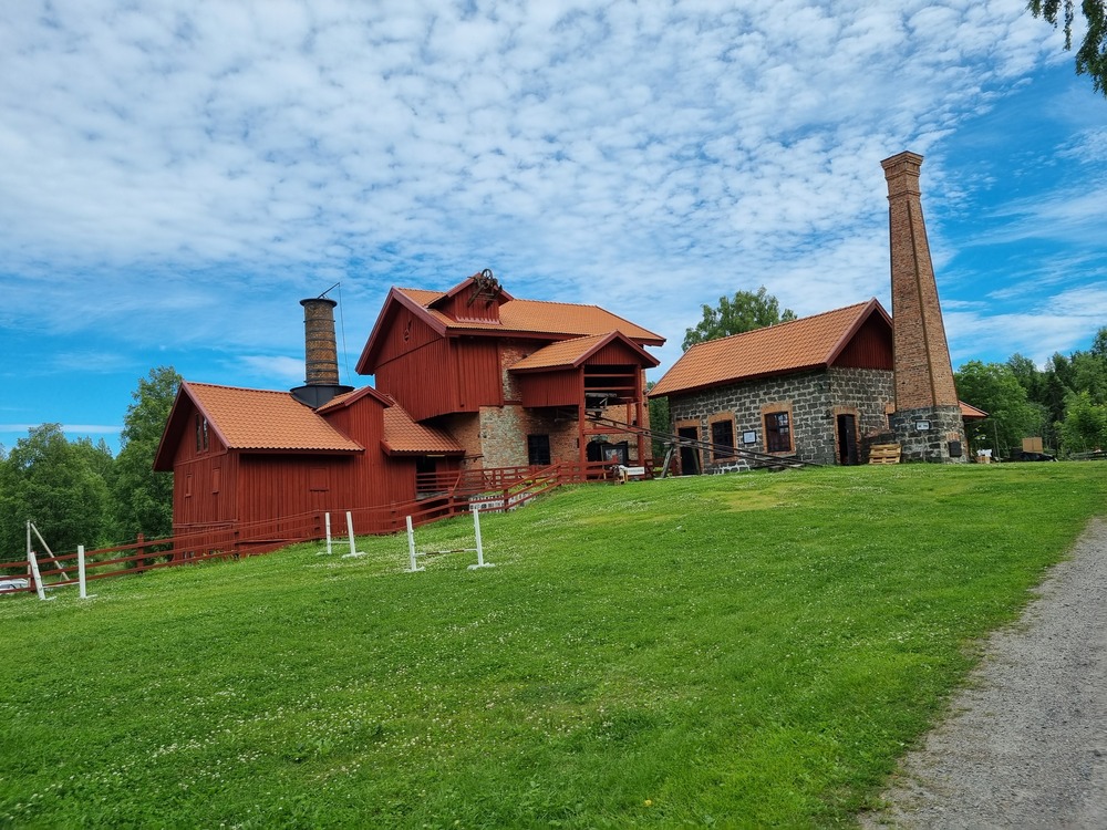 Hytta röd träbyggnad och stenhus med skorsten på en grön kulle under blå himmel med moln.