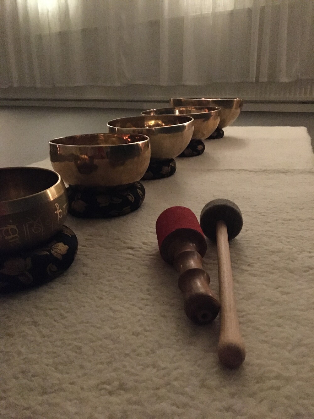 Fem tibetanska klangskålar står på rad på en matta med två klubbor framför dem i ett dämpat rum.