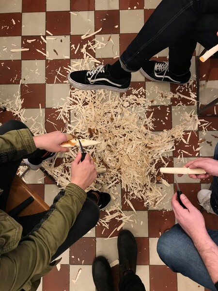 Tre personer täljer träpinnar och träspån ligger utspridda på ett rutigt golv.