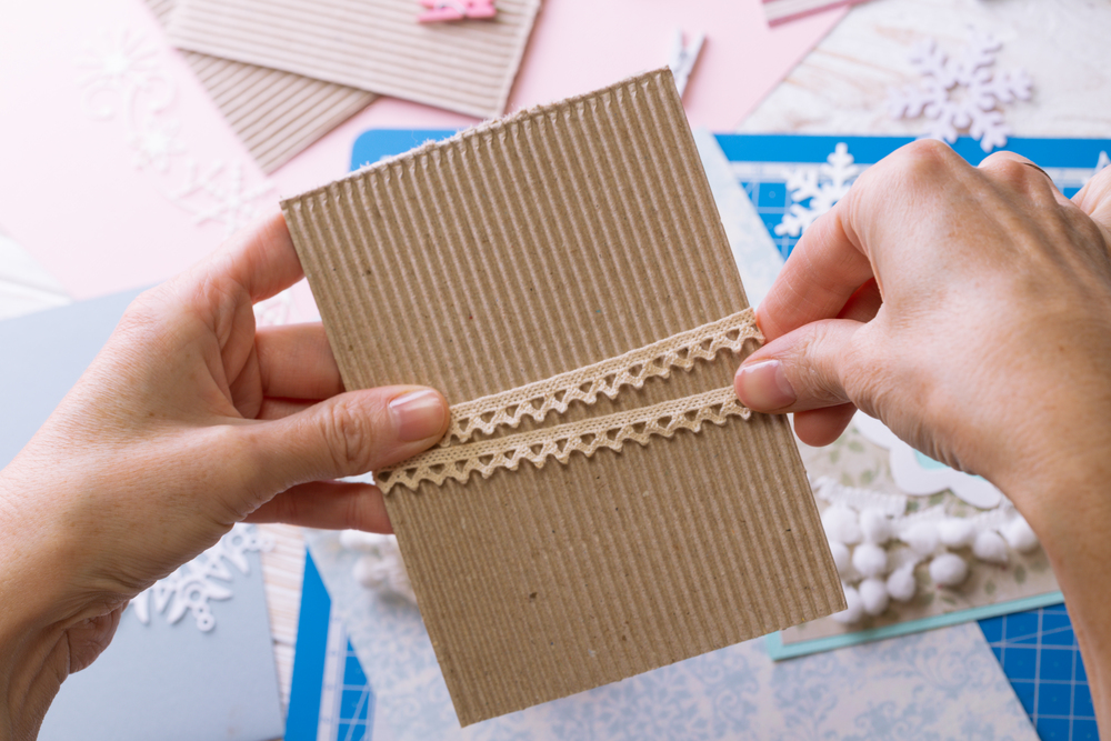 Två händer dekorerar ett kort av wellpapp med spetsband på ett pysselbord.