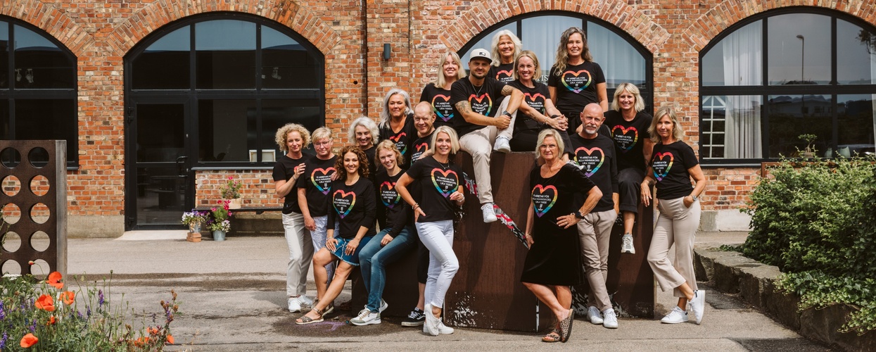 En grupp glada personer i svarta t-shirts med färgglada hjärtan poserar utomhus framför en tegelbyggnad.