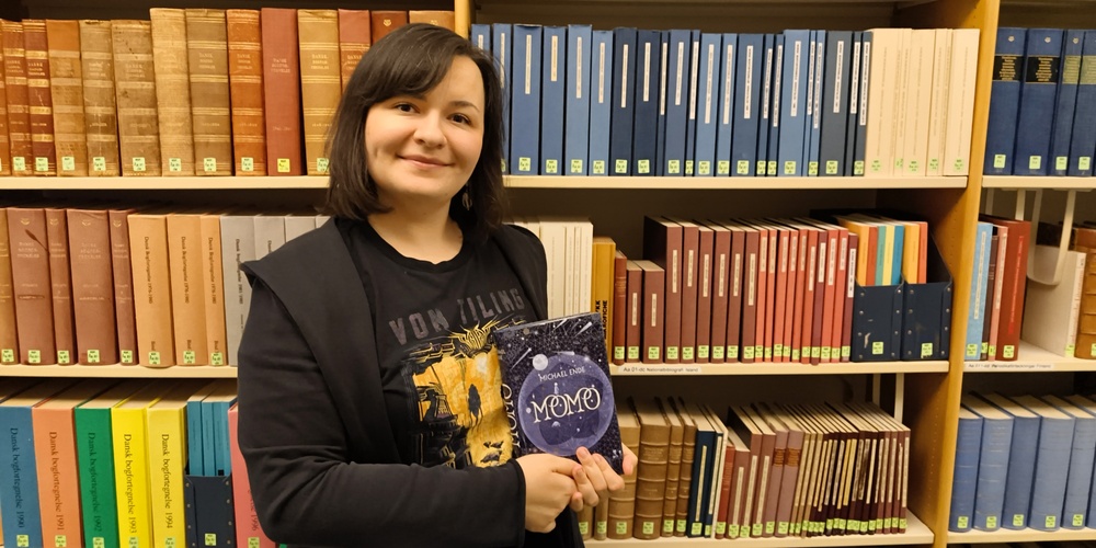 Person står i ett bibliotek och håller en bok med titeln 'Meme'.