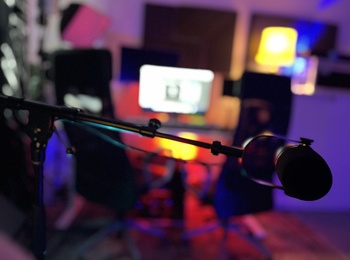 Närbild på en mikrofon i ett färgglatt, suddigt inspelningsstudio med dator i bakgrunden.