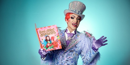 En dragqueen i färgglad kostym och hatt håller upp en barnbok mot en turkos bakgrund.