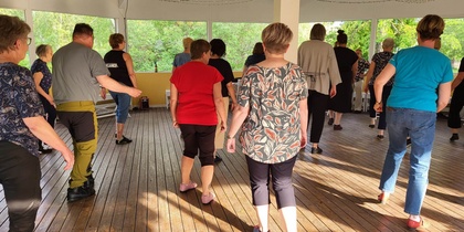En grupp vuxna dansar tillsammans på ett trägolv i en ljus paviljong med stora fönster.