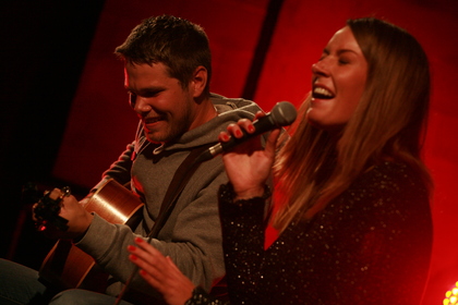 Emil spelar gitarr och Zandra sjunger i mikrofon på en scen med röd belysning.
