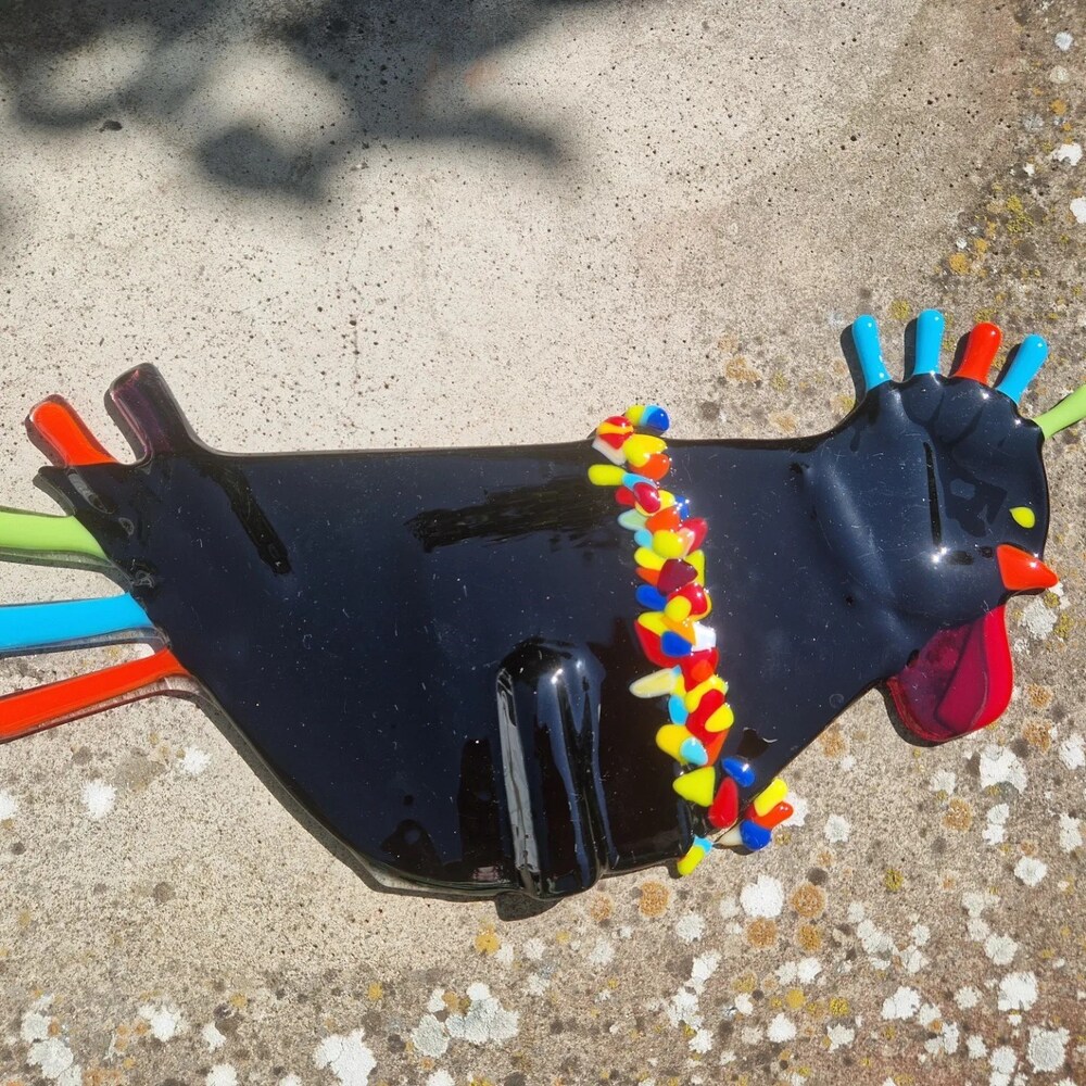 Färgglad glaskonst i form av en tupp ligger på en betongyta utomhus.