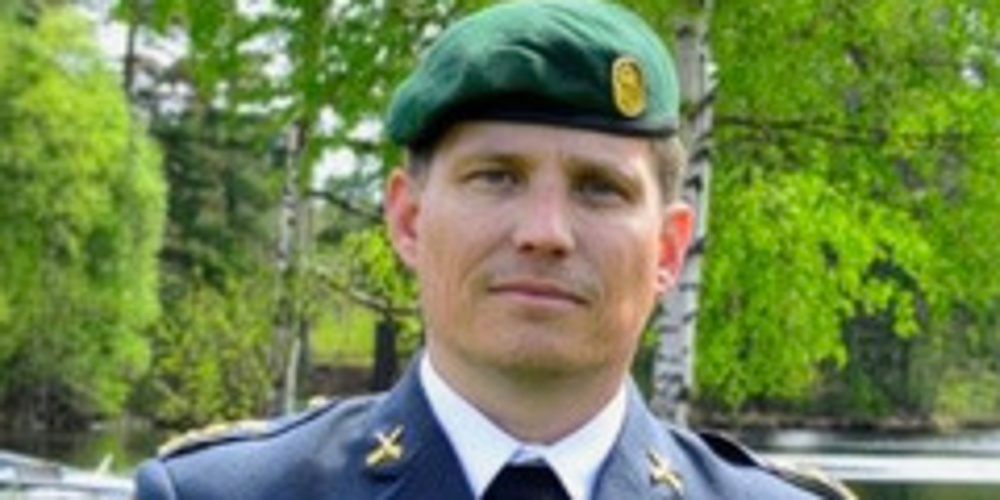 Man i militäruniform med grönt basker står utomhus framför träd och vatten.