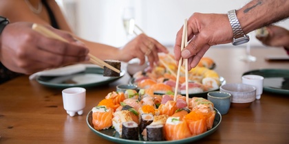 Två personer plockar sushi med ätpinnar från ett fat på ett matbord.