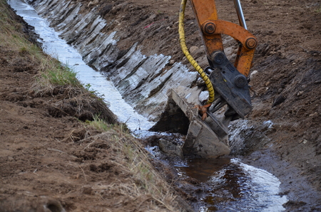 En grävmaskin gräver ett dike med vatten i en jordig miljö.