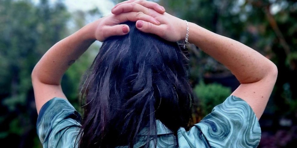 Person med mörkt hår står utomhus med ryggen mot kameran och händerna på huvudet.