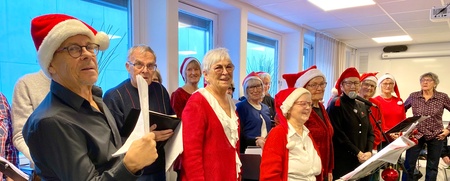 En grupp äldre personer sjunger tillsammans inomhus, många bär tomteluvor och röda kläder.