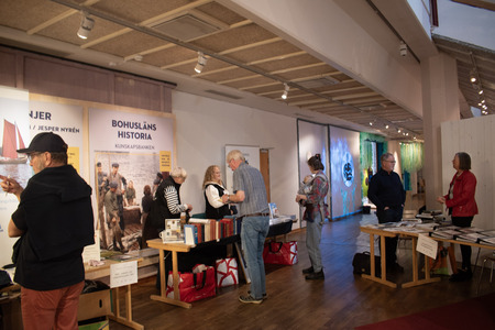 Människor samtalar och bläddrar bland böcker vid bokbord på en inomhusmässa med historietema.