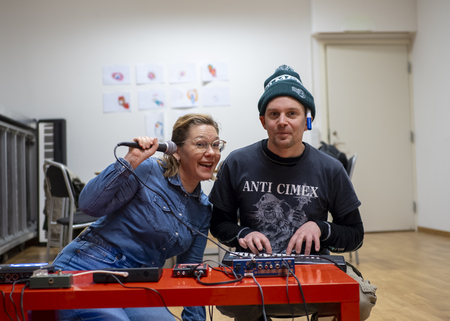 Två personer sitter vid ett rött bord med ljudutrustning, en håller en mikrofon och ler, den andra bär mössa och t-shirt.
