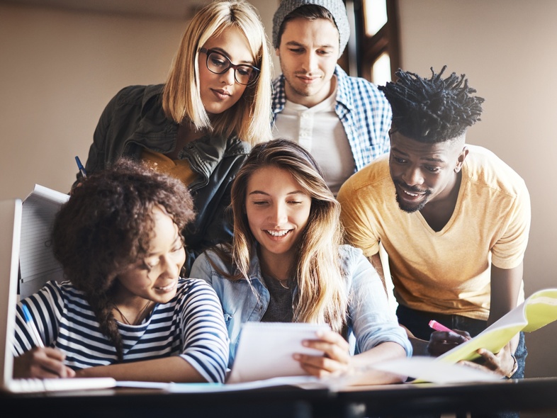 Fem unga vuxna studerar tillsammans vid ett bord, tittar på papper och ler.