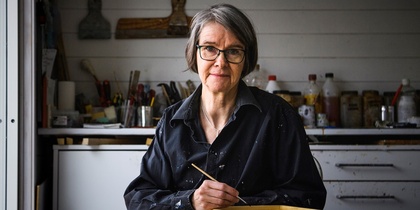 Person med glasögon och mörk skjorta sitter vid ett bord i en ateljé och målar med pensel.