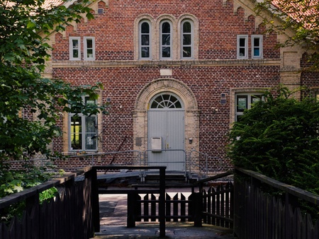 En gammal tegelbyggnad med spetsigt tak, sedd från en träbro omgiven av grönska.