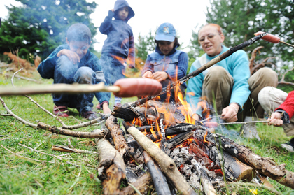 Flera personer grillar korv över öppen eld utomhus i en grön skogsmiljö.