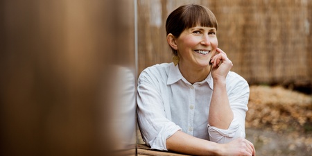 Leende kvinna med ljust skjorta sitter vid ett träbord utomhus, med handen mot kinden.