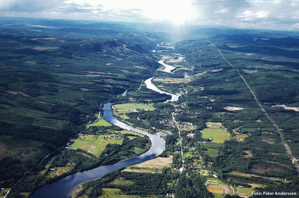 Flygfoto över en slingrande flod som rinner genom ett grönt skogslandskap med öppna fält och små samhällen.