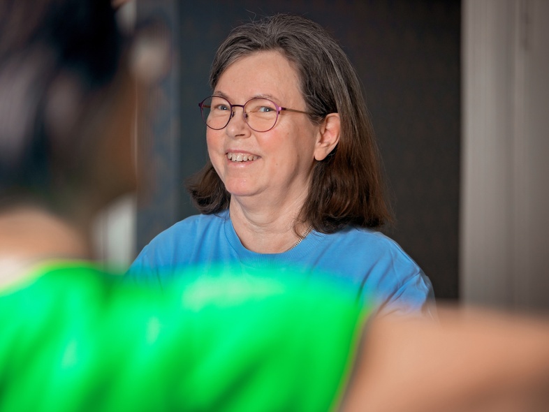 Leende kvinna med glasögon och blå tröja sitter inomhus, suddig person i förgrunden.