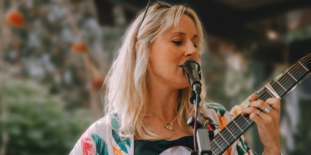 Kvinna i färgglad klänning spelar gitarr och sjunger vid en mikrofon på en utomhusscen.