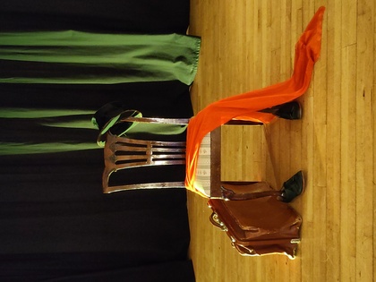 En stol på en scen med en orange sjal, svart hatt, skor och en brun portfölj, men ingen person syns.