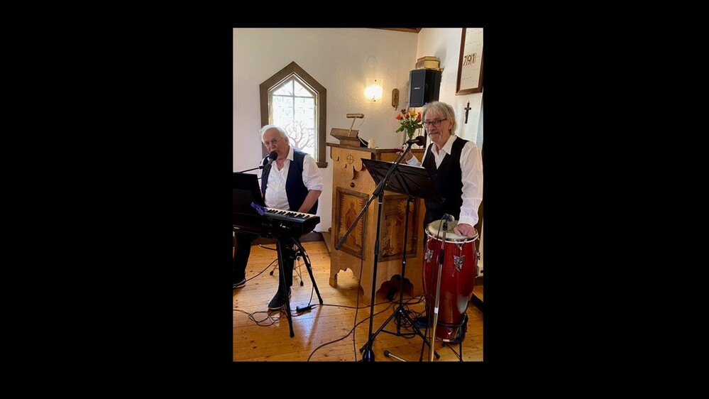 Två musiker spelar keyboard och congas i en liten kyrka, båda klädda i vit skjorta och svart väst.