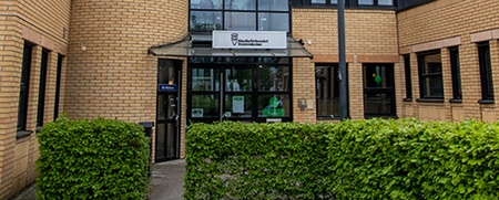 Entré till en modern byggnad med ljusgul tegelfasad, stora fönster och glasparti i mitten.