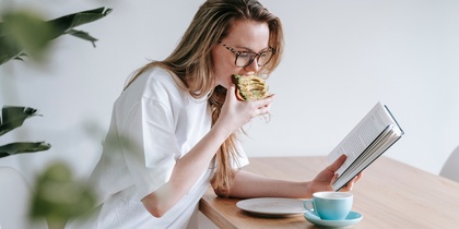 Kvinna med glasögon äter en smörgås och läser en bok vid ett träbord med en kopp kaffe.