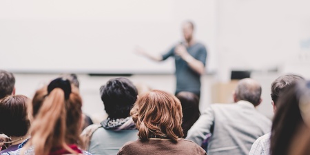 En grupp människor sitter och lyssnar på en person som håller en presentation i ett ljust rum.