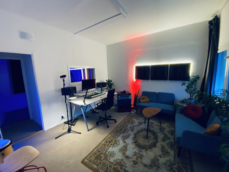 Ett modernt hemmakontor med skrivbord, datorer, mikrofon, soffa och växter, belyst med färgat ljus.