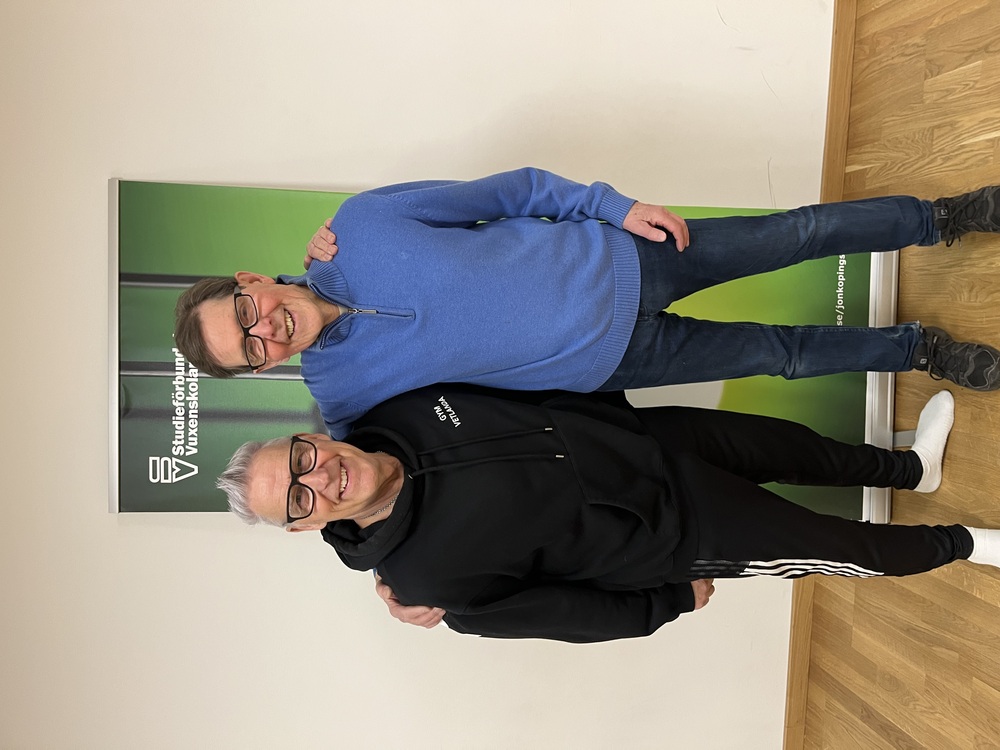 Två personer står tillsammans framför en grön skylt inomhus, en i svart hoodie och en i blå tröja.