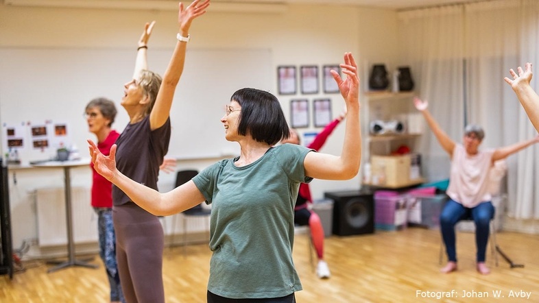 En grupp vuxna deltar i en dansklass i en ljus träningssal, med armarna uppsträckta och leenden på läpparna.