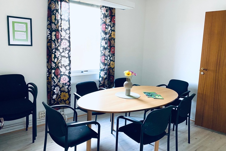 Litet mötesrum med ovalt bord, sex svarta stolar, blommig gardin och vas med blommor på bordet.