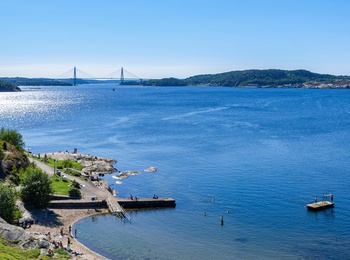 Solig badplats vid havet med gräsytor, klippor och en bro i bakgrunden.