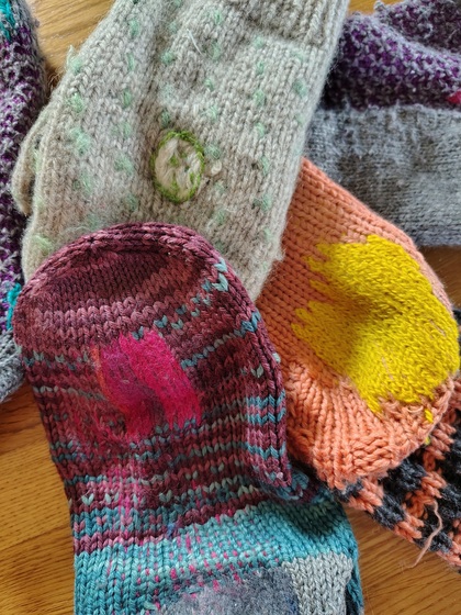 Flera stickade sockor med färgglada lagningar ligger på ett trägolv.