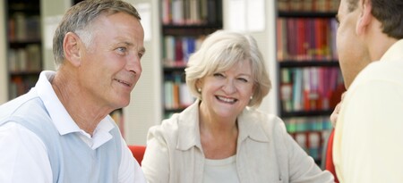 Två äldre personer samtalar med en yngre man i ett bibliotek. Alla ser glada ut.