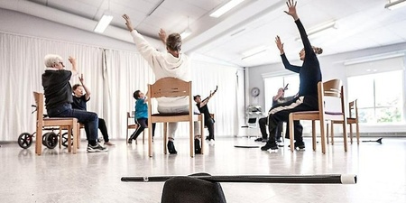 En grupp personer sitter på stolar i en ljus sal och sträcker armarna uppåt, hatt och käpp i förgrunden.